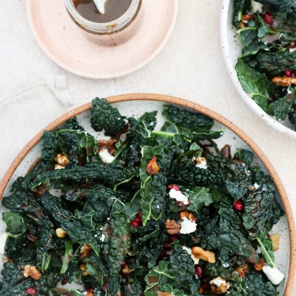 A kale and pomegranate salad is on a grey speckled plate with a brown rim. There is a small jar of balsamic dressing on the side.
