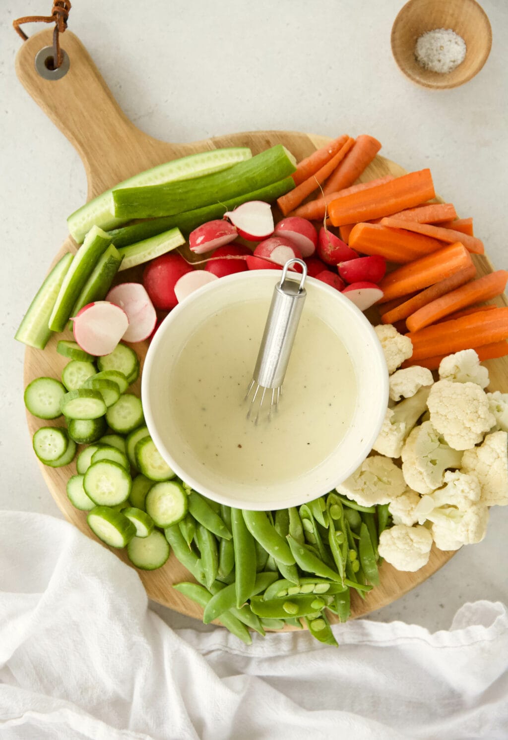 a bowl of creamy caesar salad dressing with Greek yogurt is in a white bowl on a wooden circular cutting board. THe bowl is surrounded by sliced vegetables like snap peas, cauliflower, carrots, cucumber, and radishes.