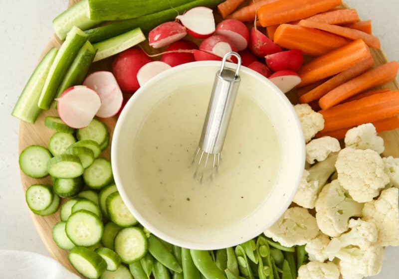 a bowl of creamy caesar salad dressing with Greek yogurt is in a white bowl on a wooden circular cutting board. THe bowl is surrounded by sliced vegetables like snap peas, cauliflower, carrots, cucumber, and radishes.