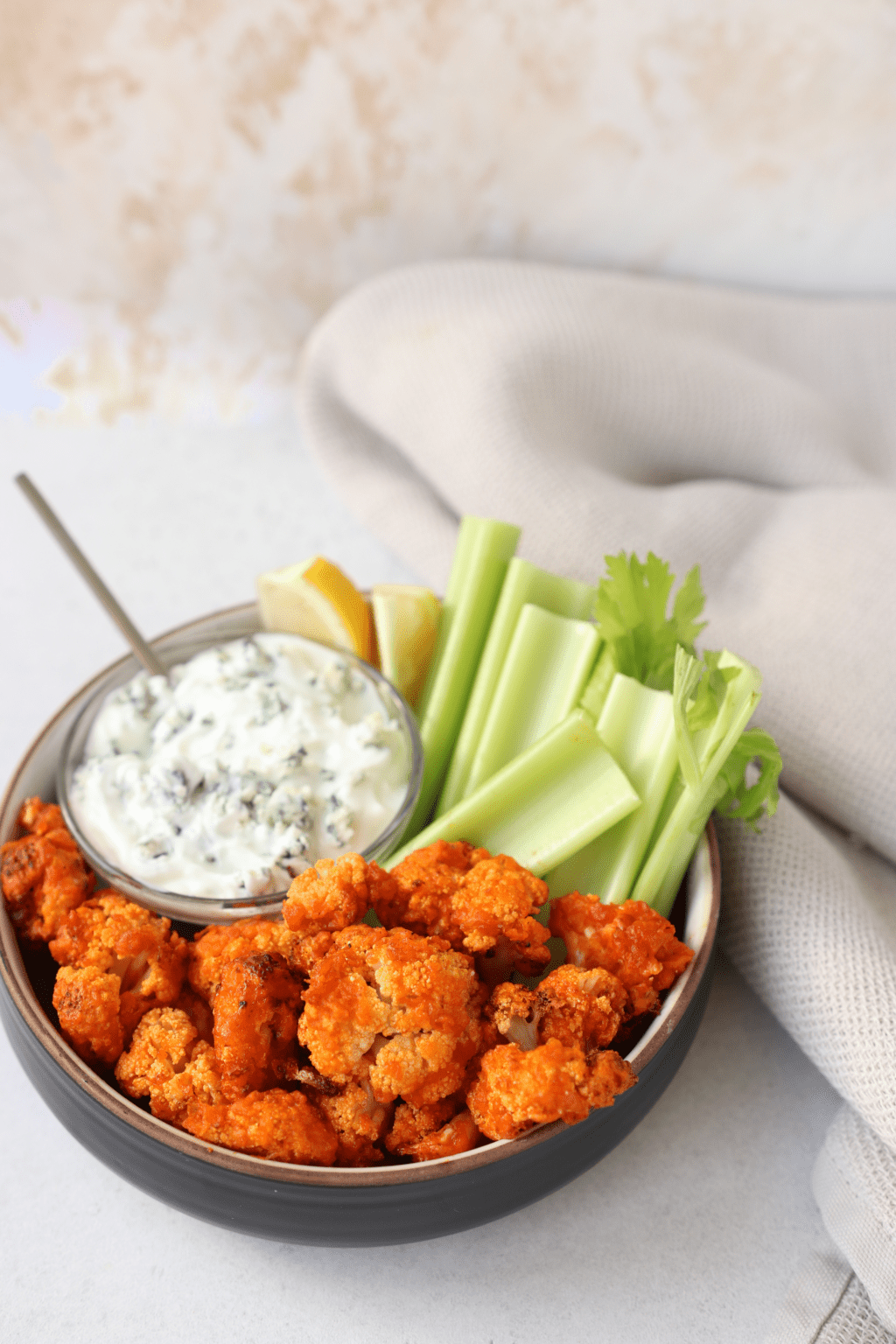 A bowl is filled with crispy buffalo cauliflower bites, celery sticks and a smaller bowl of ranch sauce with a small spoon in it.