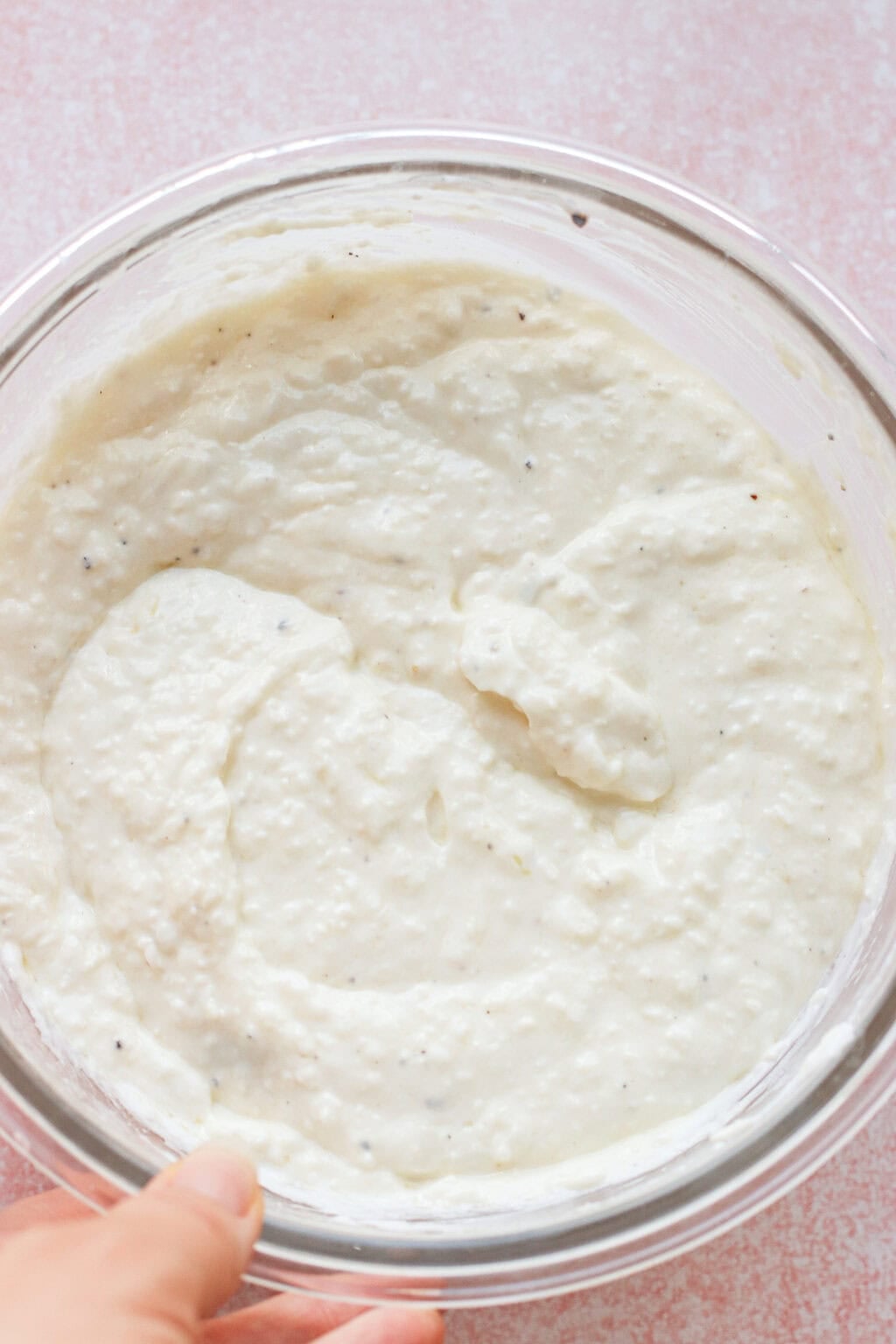 A hand holds a glass bowl filled with thick, creamy caesar dressing with greek yogurt, speckled with black pepper on a light pink surface.