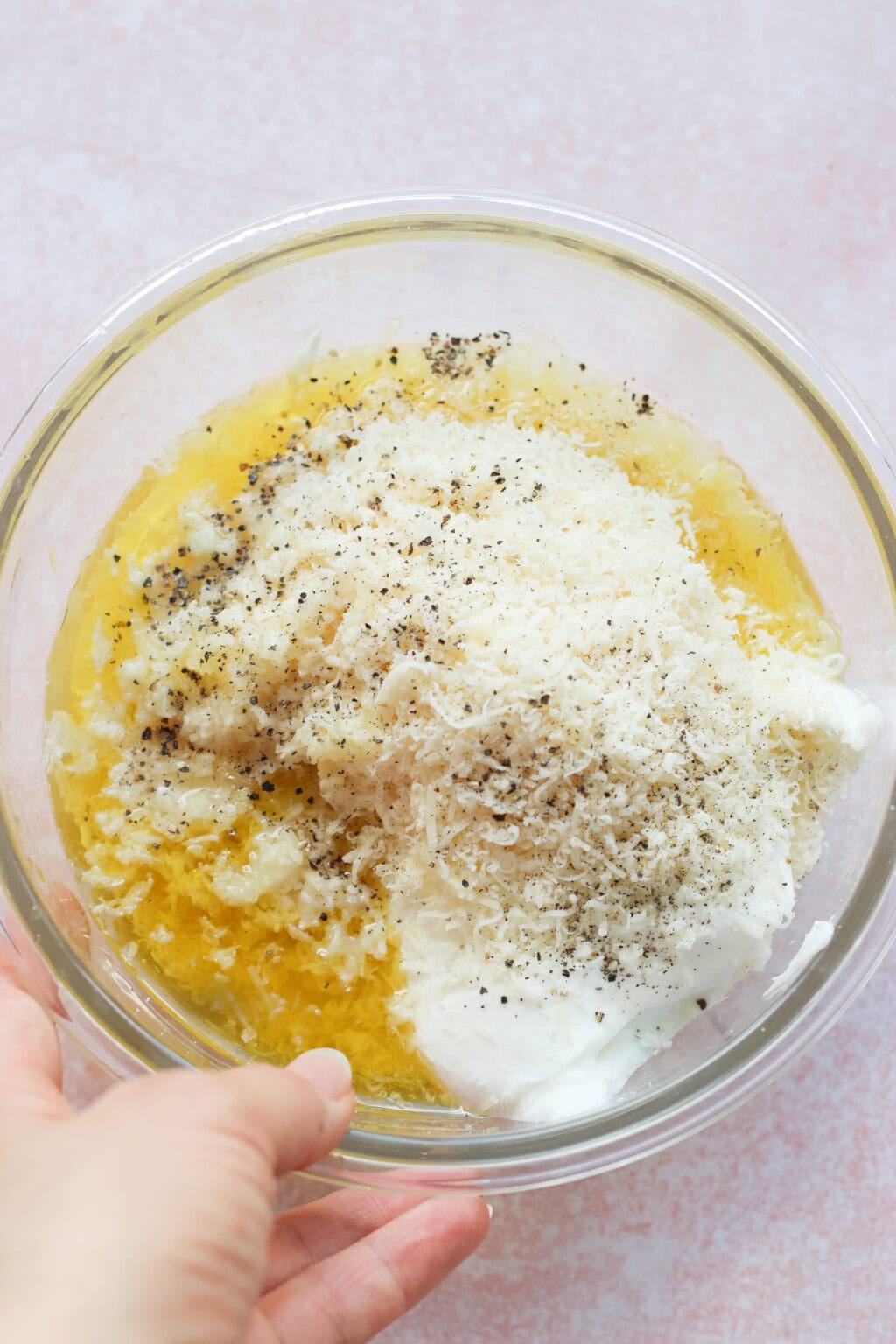 A hand holds a glass bowl filled with shredded cheese, caesar dressing with greek yogurt, beaten eggs, black pepper, and other ingredients, ready to be mixed on a light pink surface.