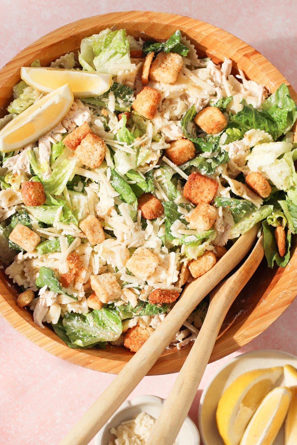 A wooden bowl filled with Caesar salad, featuring romaine lettuce, shredded chicken, croutons, grated cheese, lemon wedges, and caesar dressing with greek yogurt. Wooden tongs rest nearby with extra lemon wedges and a small dish of grated cheese.