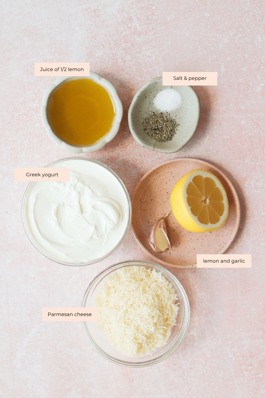 Five labeled bowls and plates on a pink surface: olive oil, salt and pepper, Greek yogurt for caesar dressing with greek yogurt, half a lemon with a garlic clove, and grated Parmesan cheese.