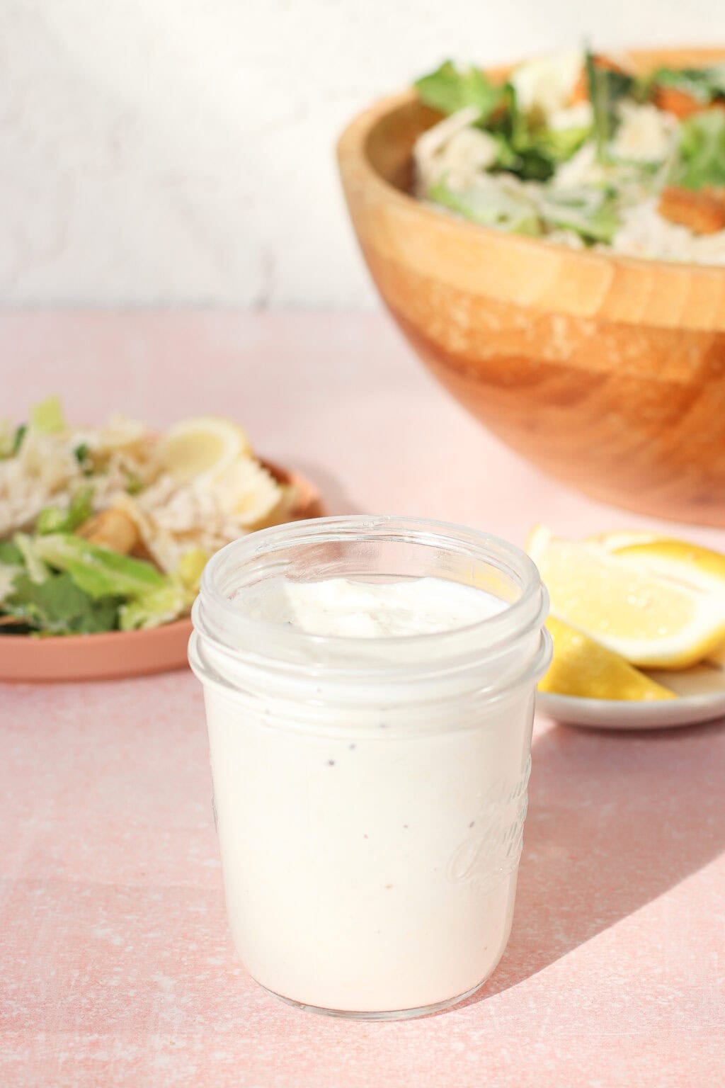 A glass jar filled with creamy white caesar dressing with greek yogurt sits on a pink surface. In the background, there’s a wooden bowl of salad, a plate of salad, and lemon wedges on a small dish.