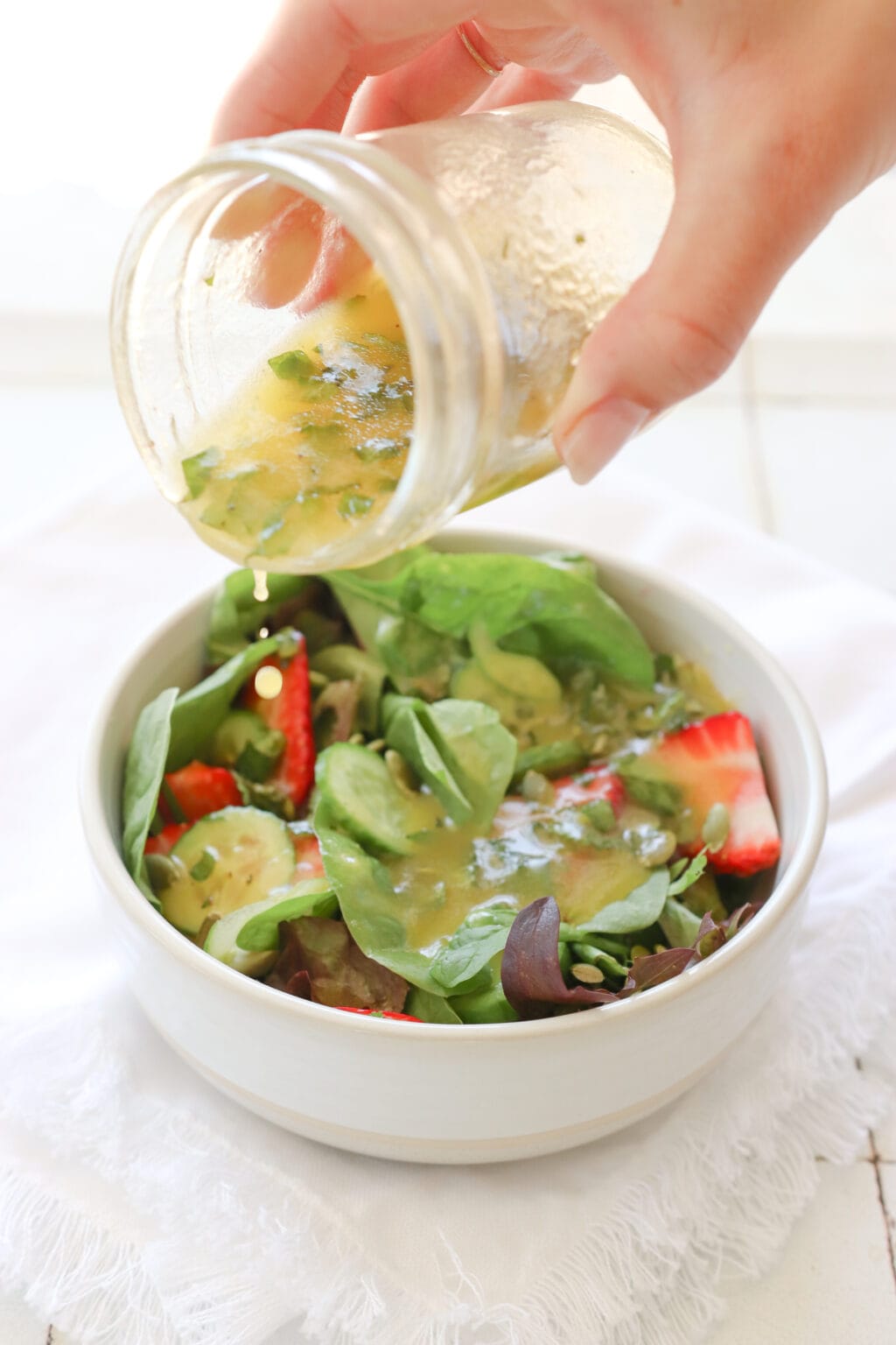 A hand holding a glass mason jar with mango salad dressing is pouring vinaigrette over a mixed green and strawberry salad. 