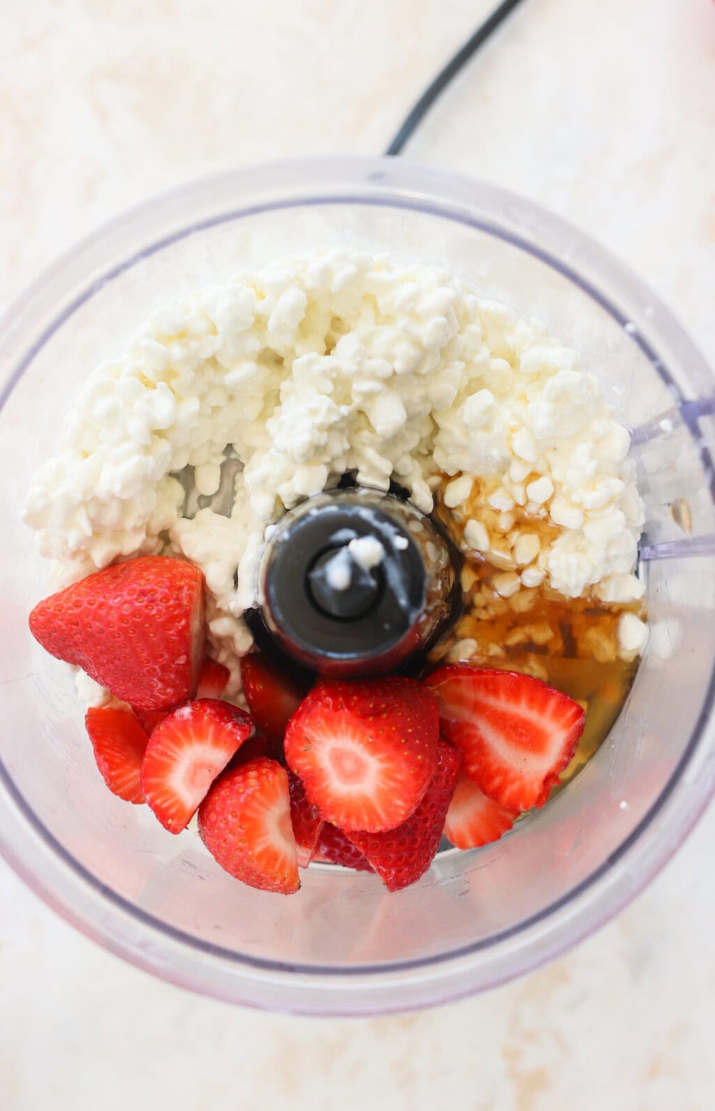 strawberries, cottage cheese and honey sit in an open food processors sitting on a white an pink mottled counter top