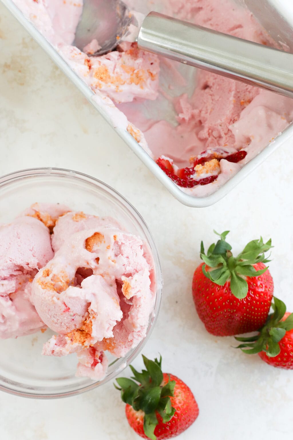 An overhead photo of pink ice cream in a bowl and in a loaf pan. There are two scoops of ice cream in a bowl, two strawberries on the countertop, and a loaf pan with a few scoops taken out of it. 