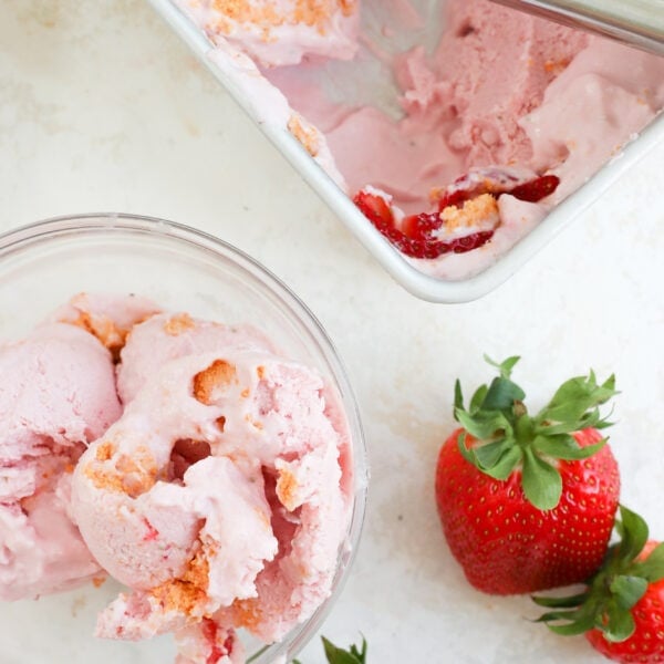 An overhead photo of strawberry cottage cheese ice cream in a bowl and in a loaf pan. There are two scoops of ice cream in a bowl, two strawberries on the countertop, and a loaf pan with a few scoops taken out of it.