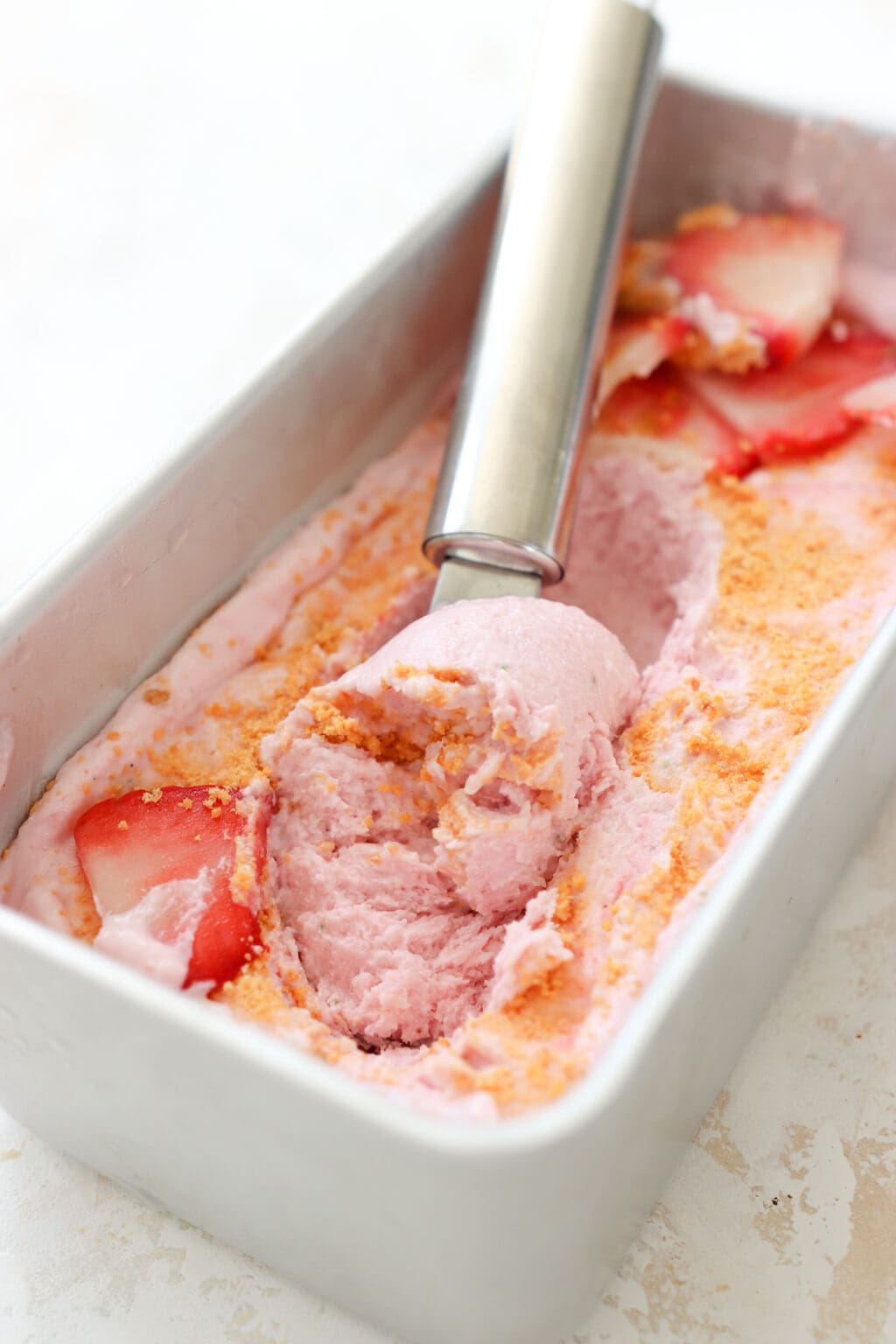 A silver ice cream scooper is scooping up some pink strawberry cottage cheese ice cream that's in a mini loaf pan. There is a graham cracker crust and sliced strawberries on the top
