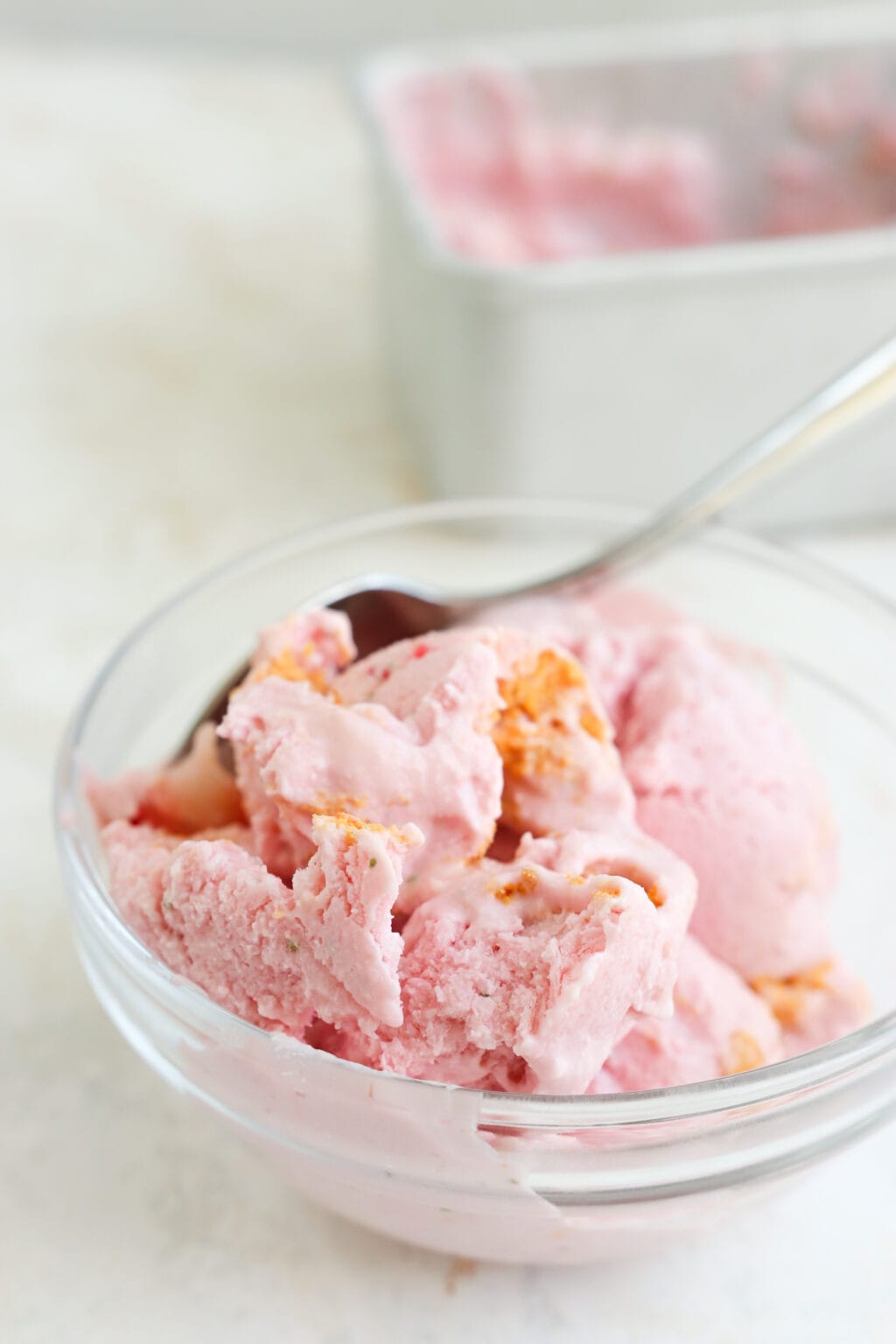 A bowl of pink strawberry cottage cheese ice cream with a silver spoon in it. In the background is a loaf pan with some cottage cheese ice cream in it.