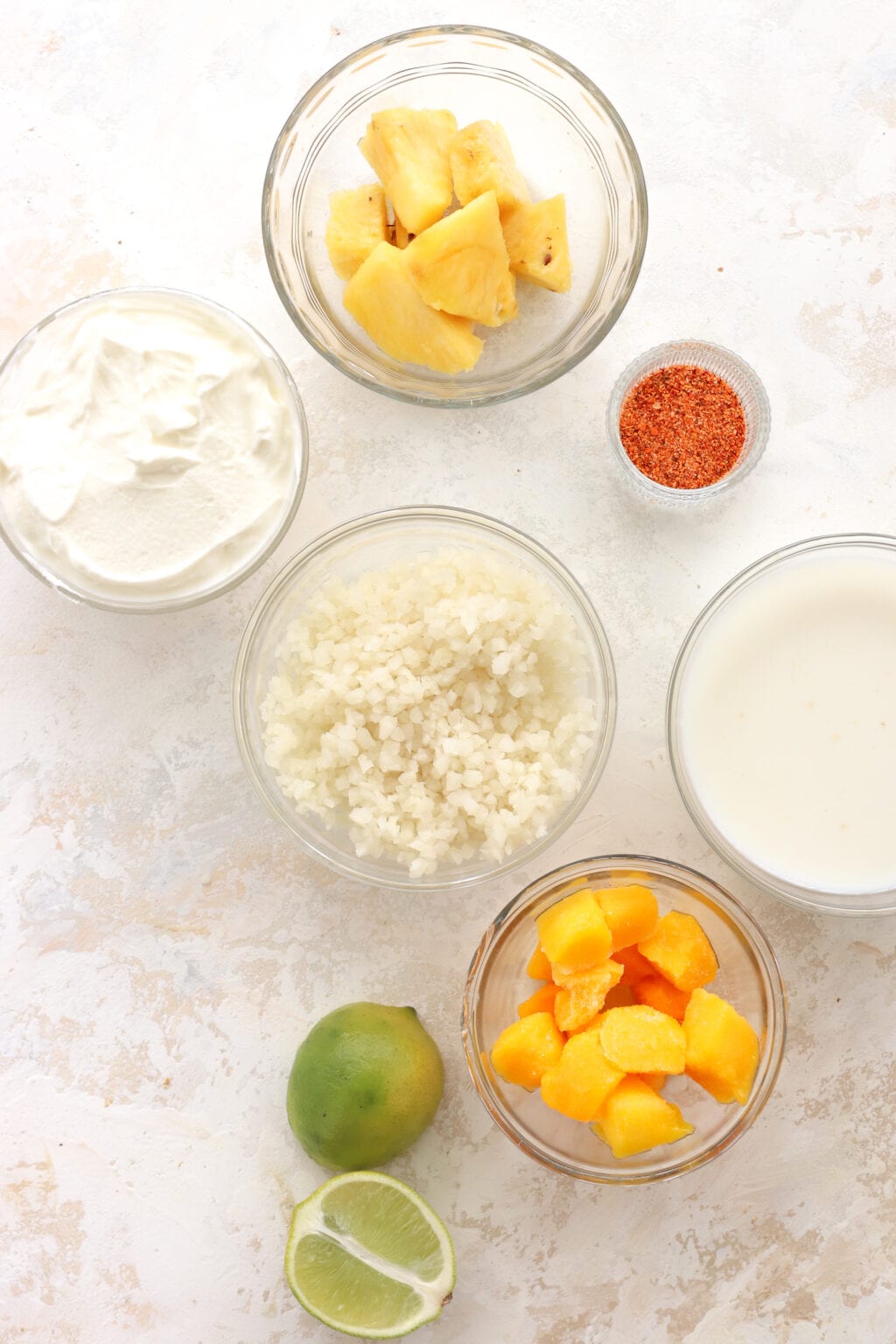 The ingredients of the mango and pineapple smoothie are laid out individually in clear bowls. At the top is a bowl of pineapple, in the middle there is a bowl of Greek yogurt, a small bowl of tajin spice, a bowl of riced cauliflower, and a bowl of milk. At the bottom is a lime cut in half beside a bowl of frozen mango.
