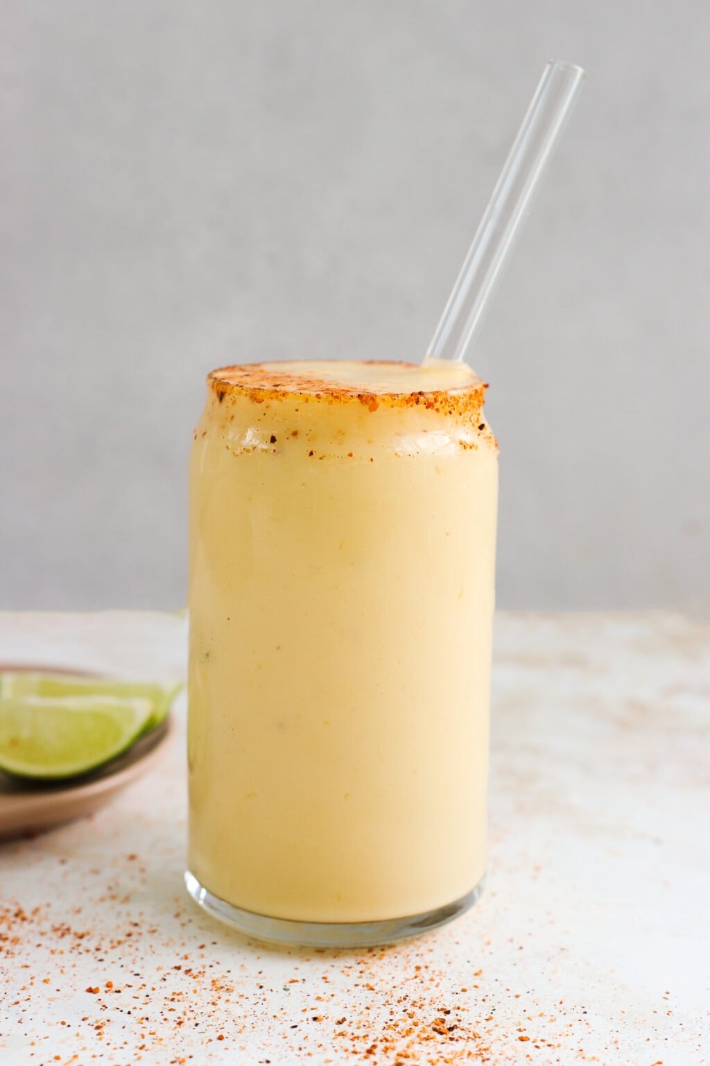 A tajin rimmed glass is filled with an orange colored mango and pineapple smoothie. There is a clear glass straw in the cup. On the counter there is a small plate with lime slices and a sprinkling of tajin spice. 