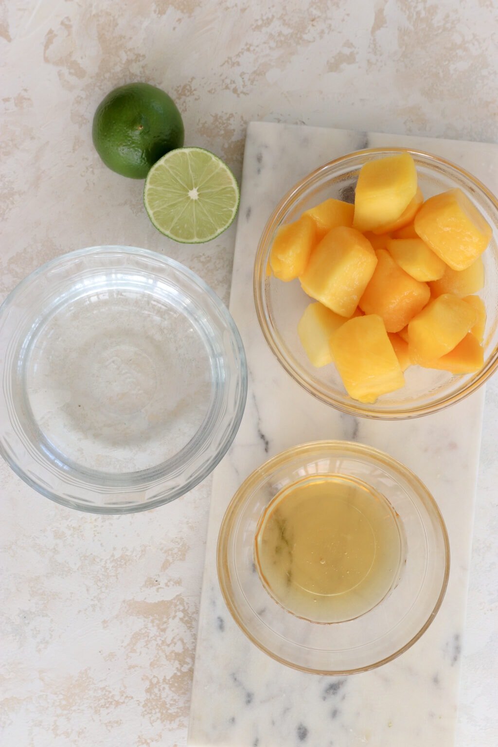 An overhead shot of ingredients for a recipe. In the top left is two lime halves, to the right of the limes is a clear bowl with cubed mango. To the lower left of the mango is a bowl of clear water. To the lower right of the water is a bowl of honey.