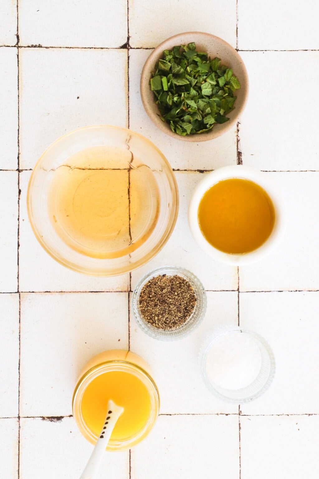An overhead shot of the ingredients for this mango and mint salad dressing. At the top is a bowl of chopped mint, c in a clear bowl is olive oil, to the right in a white bowl is white wine vinegar, in two small glass bowls is salt and pepper and at the bottom is a jar of mango puree.