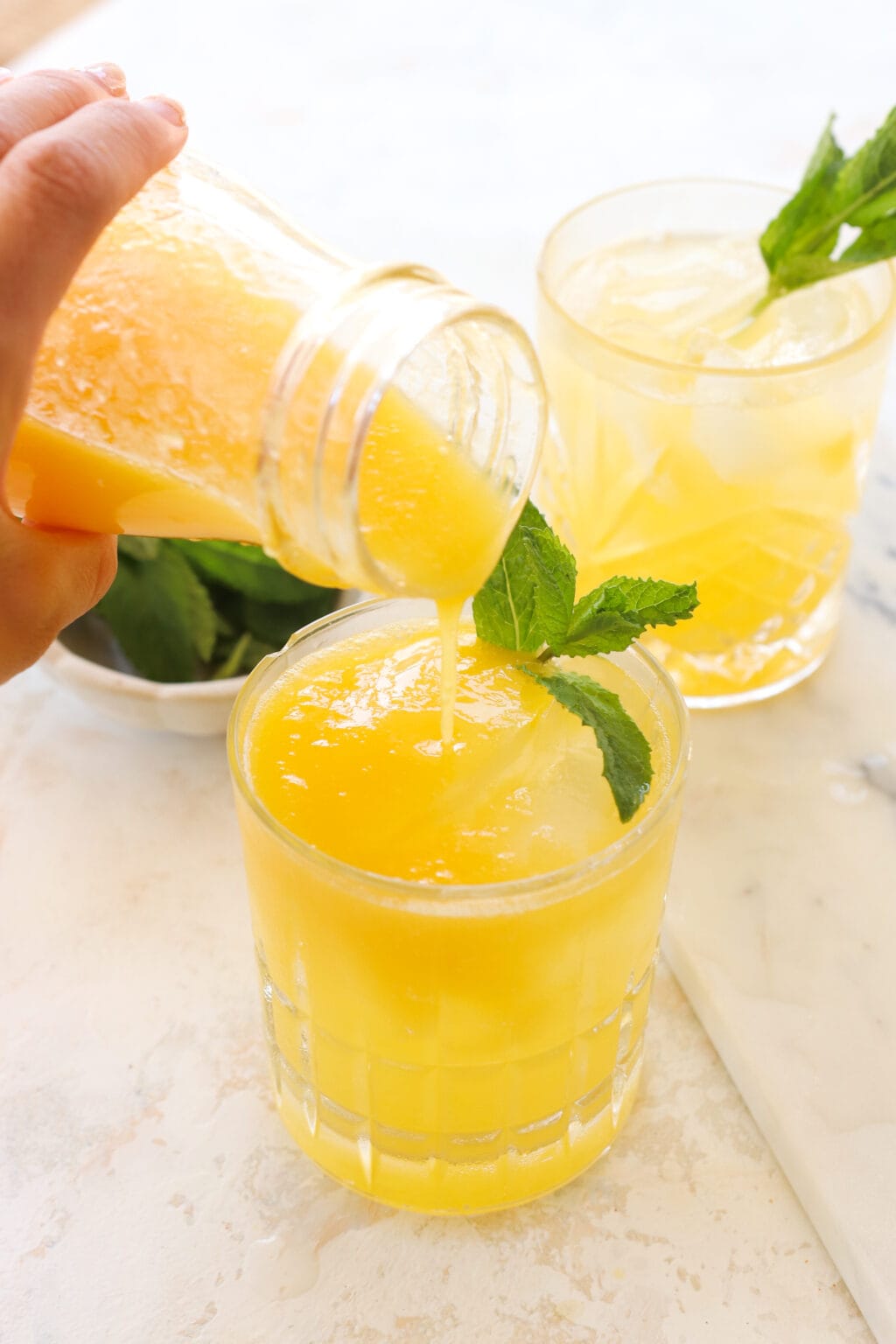 A hand holding a bottle over a short clear glass. The bottle is pouring yellowish orange puree into it and there is mint garnishing the glass. In the background is another cup with the same liquid and a bowl of mint.