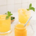 2 glasses are in front of a white tile wall. The glasses have yellowish-orange liquid in them and mint garnishes. In front of them is a smaller jar with a yellowish-orange puree and a white spoon in the jar.