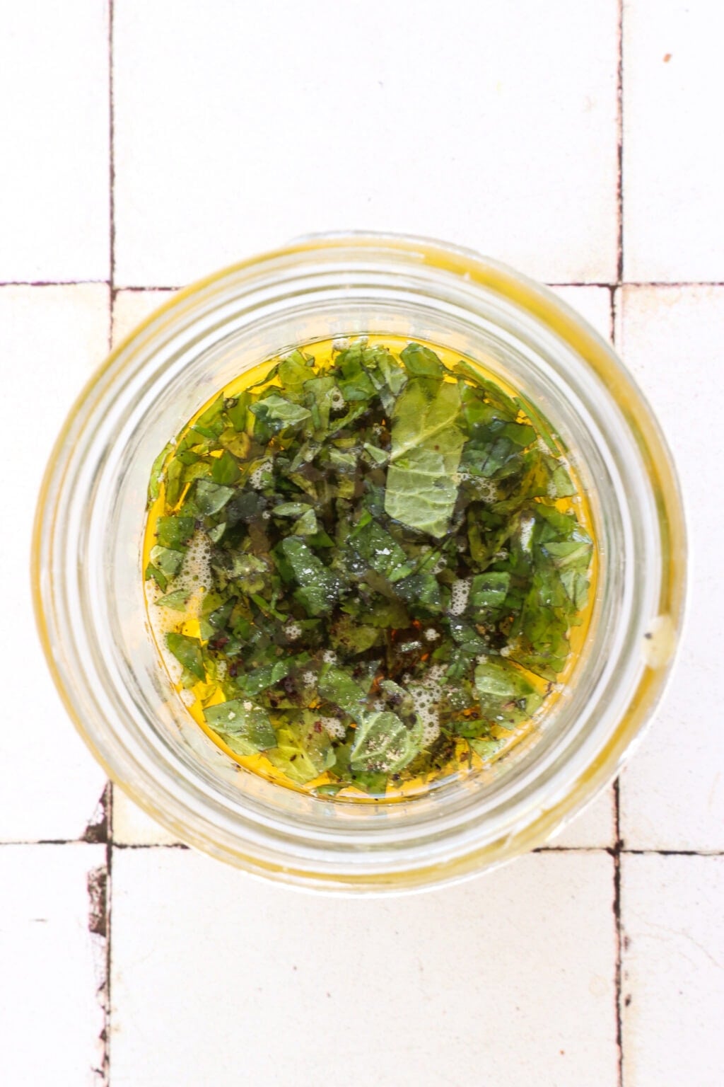 A glass bowl sits on a white tile counter. In the overhead shot is a bowl of olive oil, vinegar, mango puree, and it's topped with minced mint.