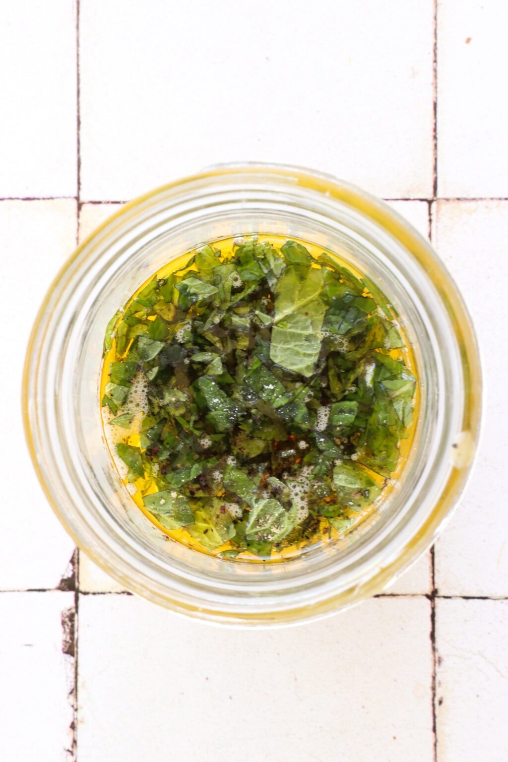 A glass bowl sits on a white tile counter. In the overhead shot is a bowl of olive oil, vinegar, mango puree, and it's topped with minced mint. 
