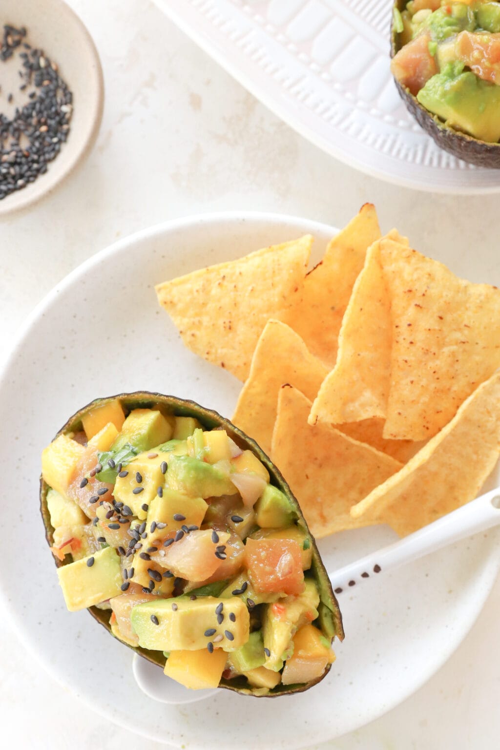 An avocado skin filled with cubed avocado, tuna and mango habanero salsa is on a white plate. There is a white spoon under the avocado and beside it are some chips.