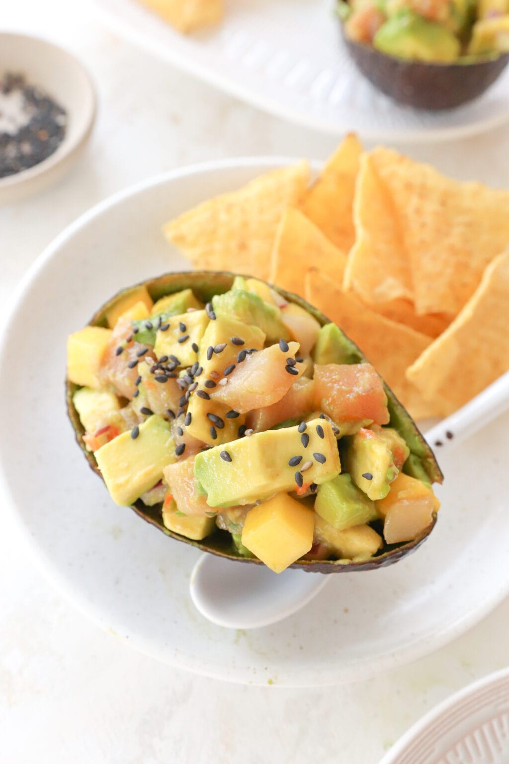 A white plate with an avocado shell filled with cubed avocado, mango and habanero salsa, and cubed tuna. Beside it are some tortilla chips and a white spoon.