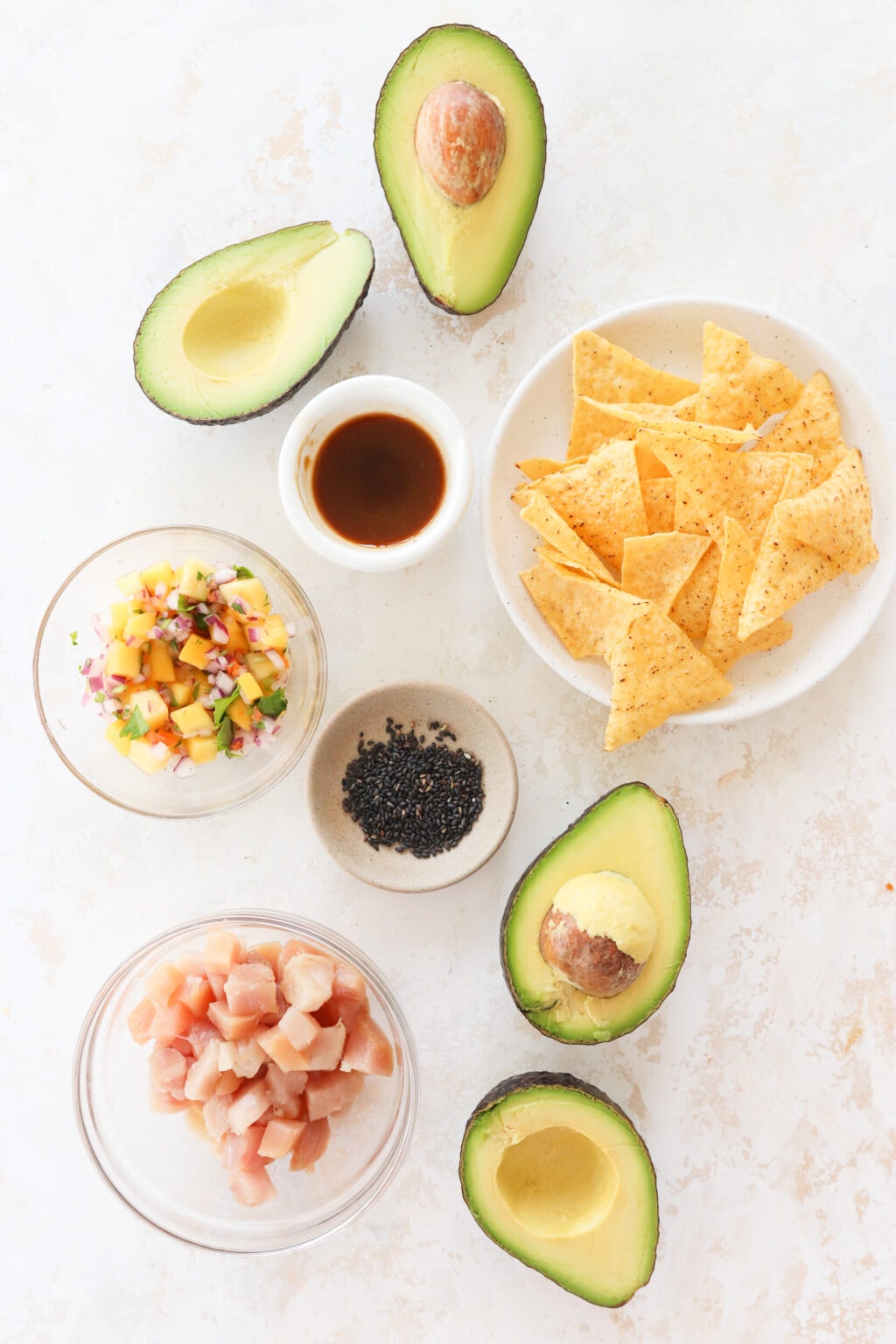 An overhead shot of ingredients for tuna avocado boats. There is an avocado cut in half, a small bowl with soy sauce, a plate of tortilla chips, a clear bowl with mango habanero salsa, a brown bowl with black sesame seeds, a clear bowl with tuna and another avocado cut into half.