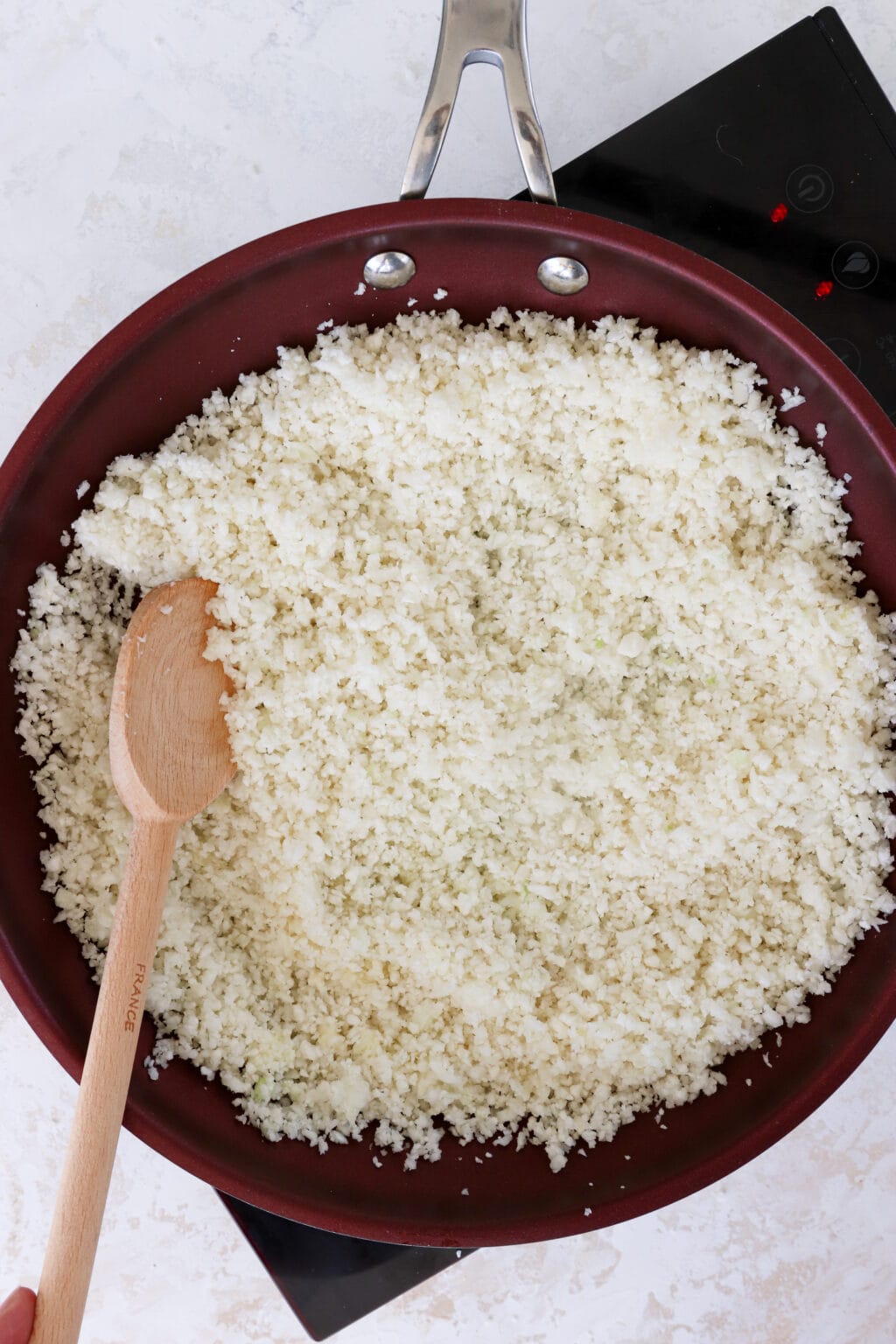 A red frying pan is fulled with riced cauliflower and bring stirred by a wooden spoon
