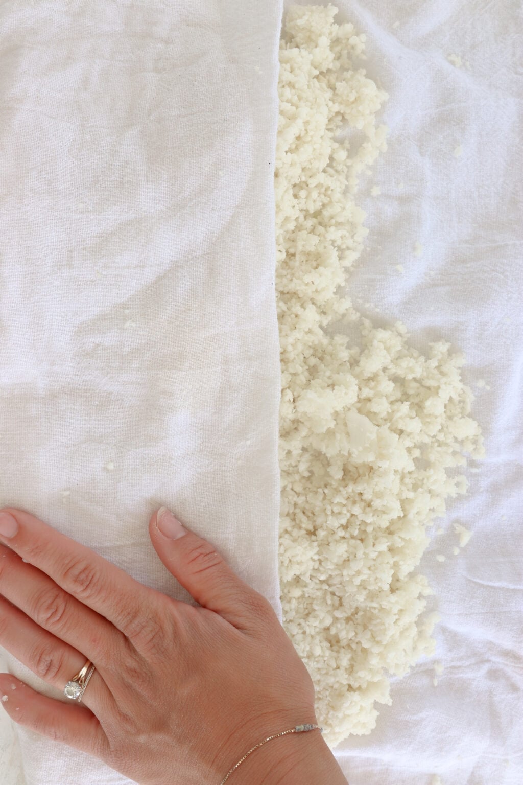 Cauliflower rice is on a white towel. There is a hand over the towel pressing the liquid out.