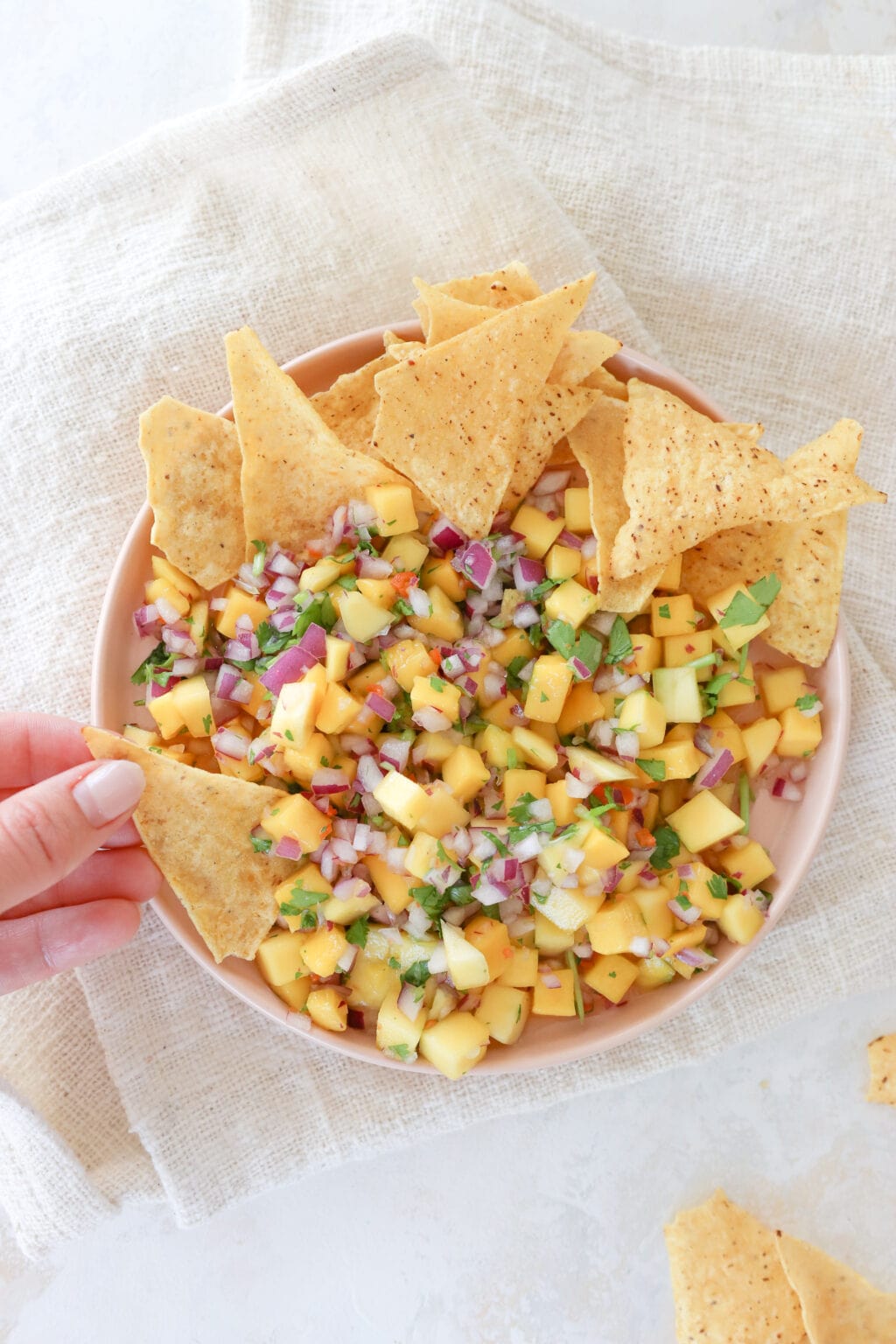 An overhead shot of a pink plate with a rim on top of a beige burlap style napkin. On the plate is a mixed mango salsa that has cubed mango, minced red onion, minced habanero pepper, and cilantro leaves. At the top of the plate is a stack on tortilla chips. A woman's hand is holding a chip and dipping it in the salsa.