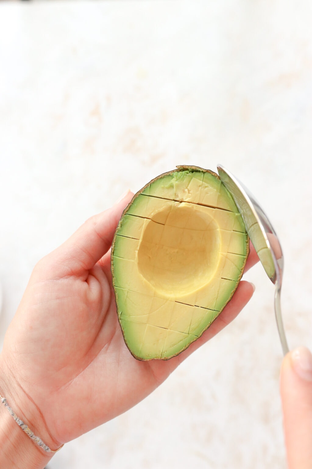 A hand is holding a scored avocado with the skin on and is using a silver spoon to scoop out the rest of the avocado. 