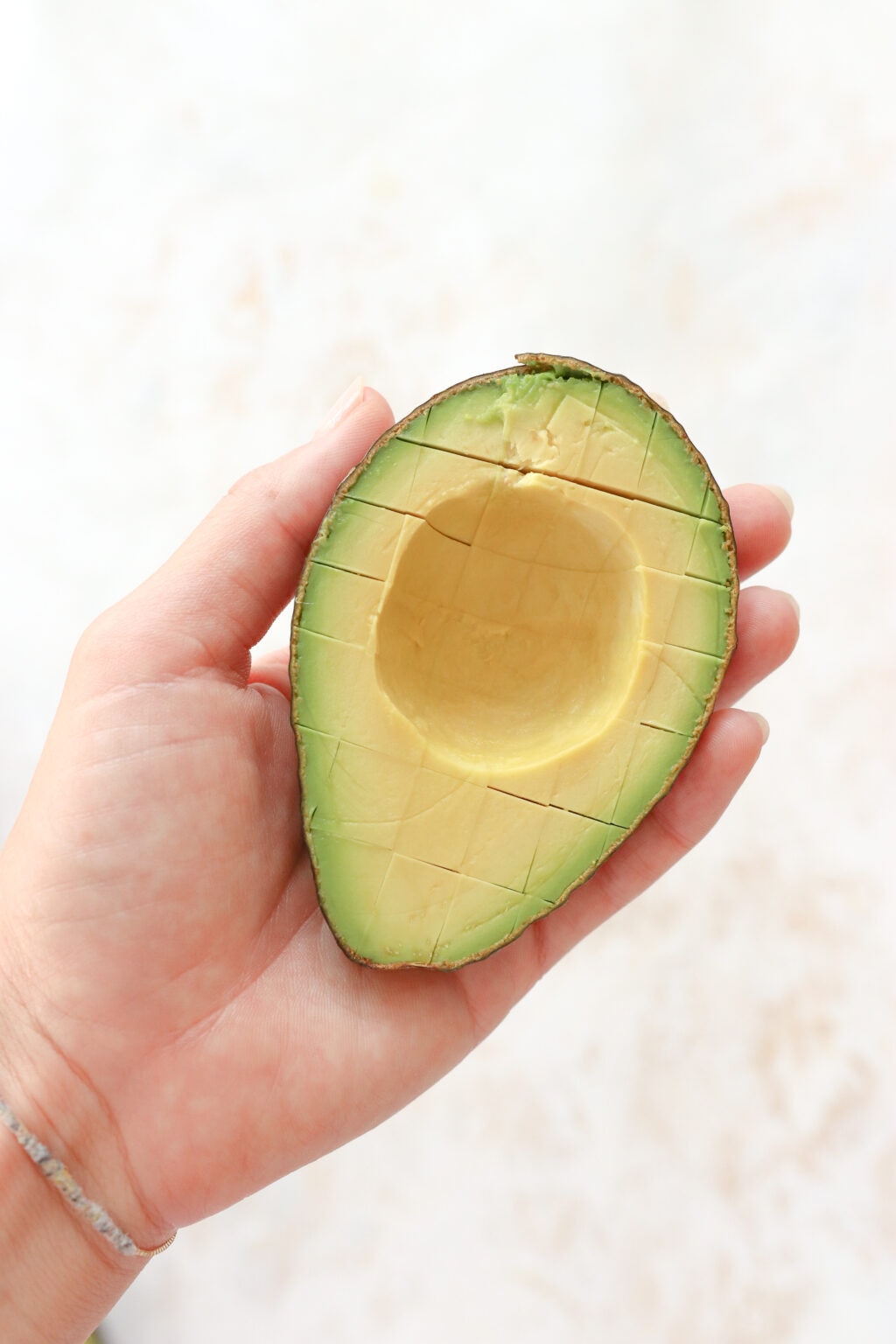 A hand is holding half an avocado. The avocado half has its skin on and has been sliced into 1/2 inch cubes.