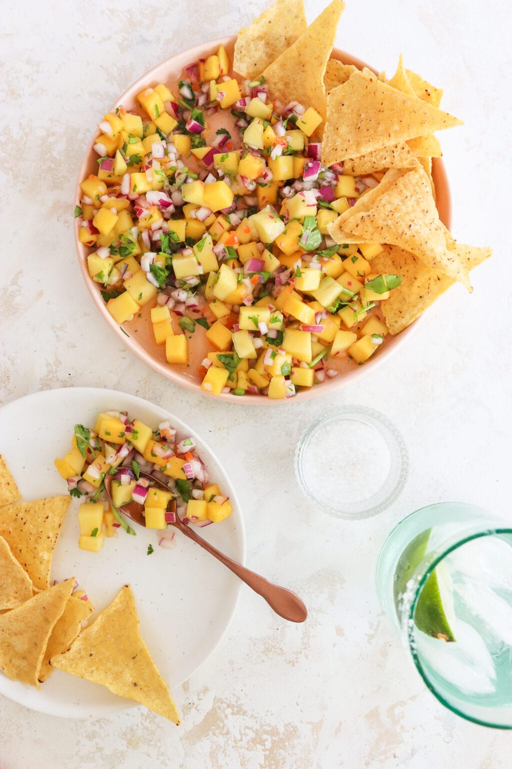 An overhead shot of a white counter top with a pink plate with a rim topped with salsa and chips. In the bottom left corner is a white plate with a copper spoon holding mango salsa and tortilla chips. In the bottom right corner is a greenish cup with soda water and lime.