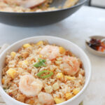 A white bowl is filled with fried rice. There is diced mango and pink prawns. The dish is topped with green onion. In the background is a bowl of chili oil and the wok of cooked fried cauliflower rice.