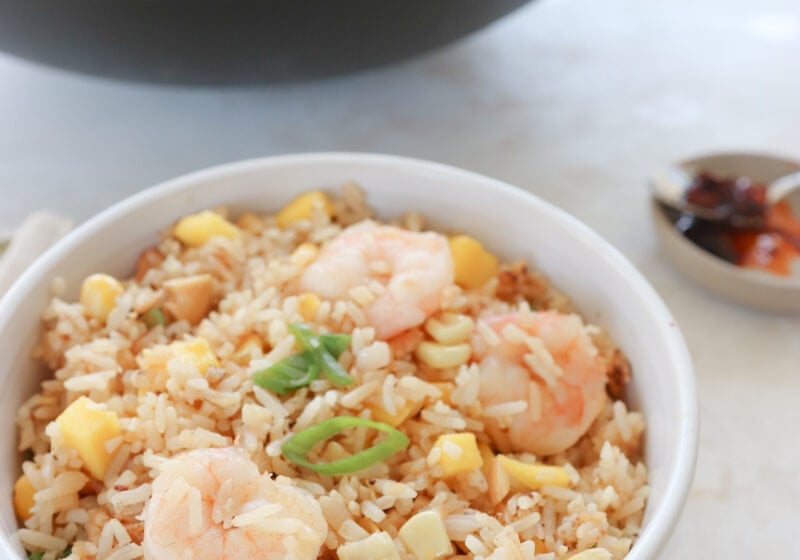 A white bowl is filled with fried rice. There is diced mango and pink prawns. The dish is topped with green onion. In the background is a bowl of chili oil and the wok of cooked fried cauliflower rice.