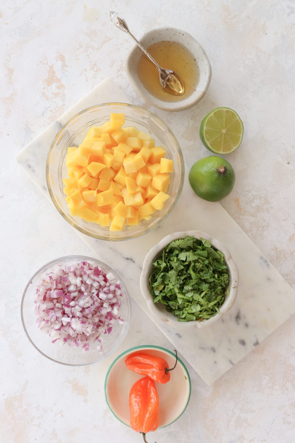 An overhead shot of ingredients to make mango habanero salsa. At the top in a grey dish is honey, below is a clear bowl with diced mango and two halves of lime. Below to the left is a small clear bowl with minced red onion and to the right is roughly chopped cilantro. At the bottom is a clear bowl with whole orange habanero peppers.  