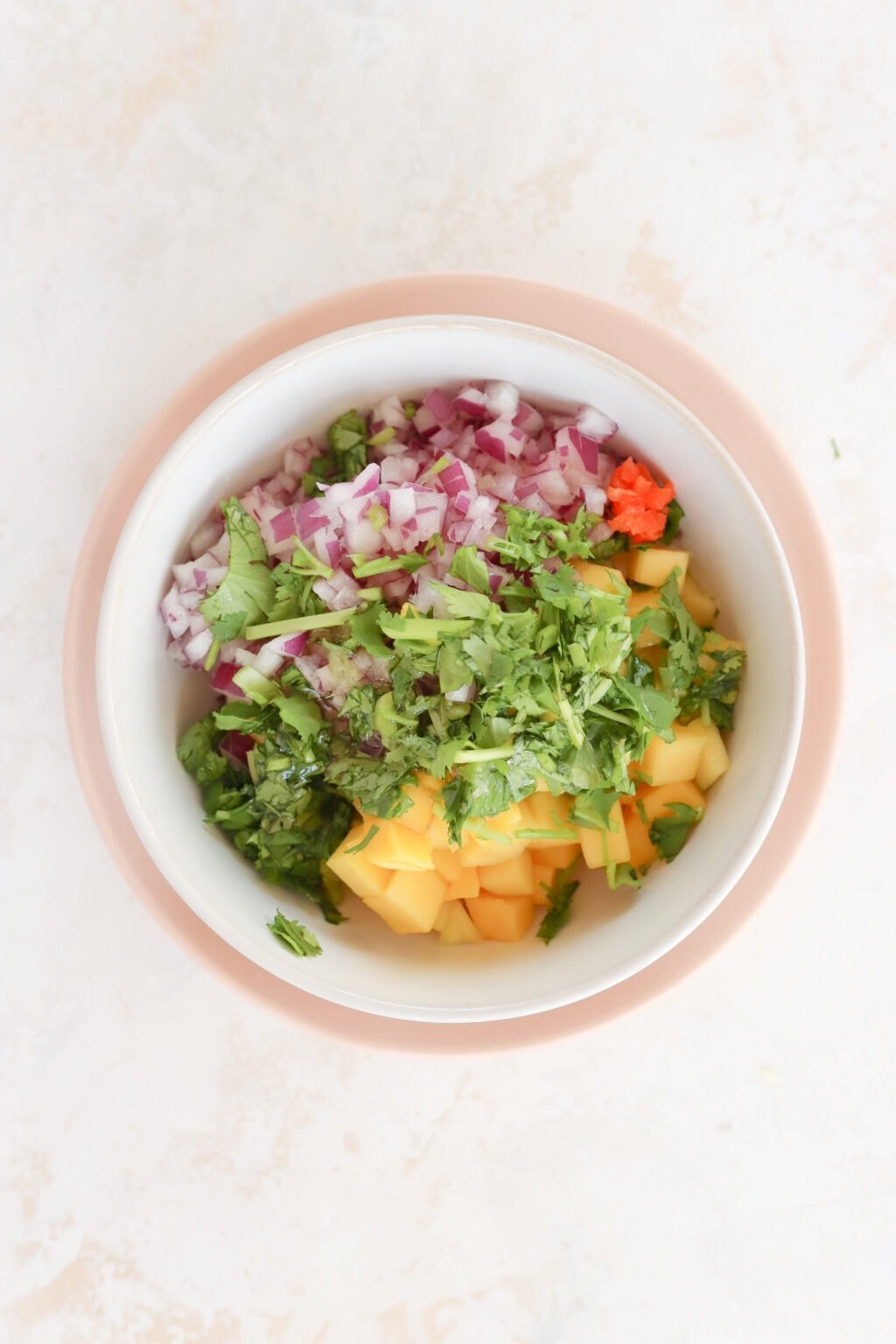 In a white bowl with a pink rim is ingredients for a salsa. There is cubed mango, diced red onion, diced habanero pepper, honey and cilantro all piled. The ingredients have not yet been mixed together.