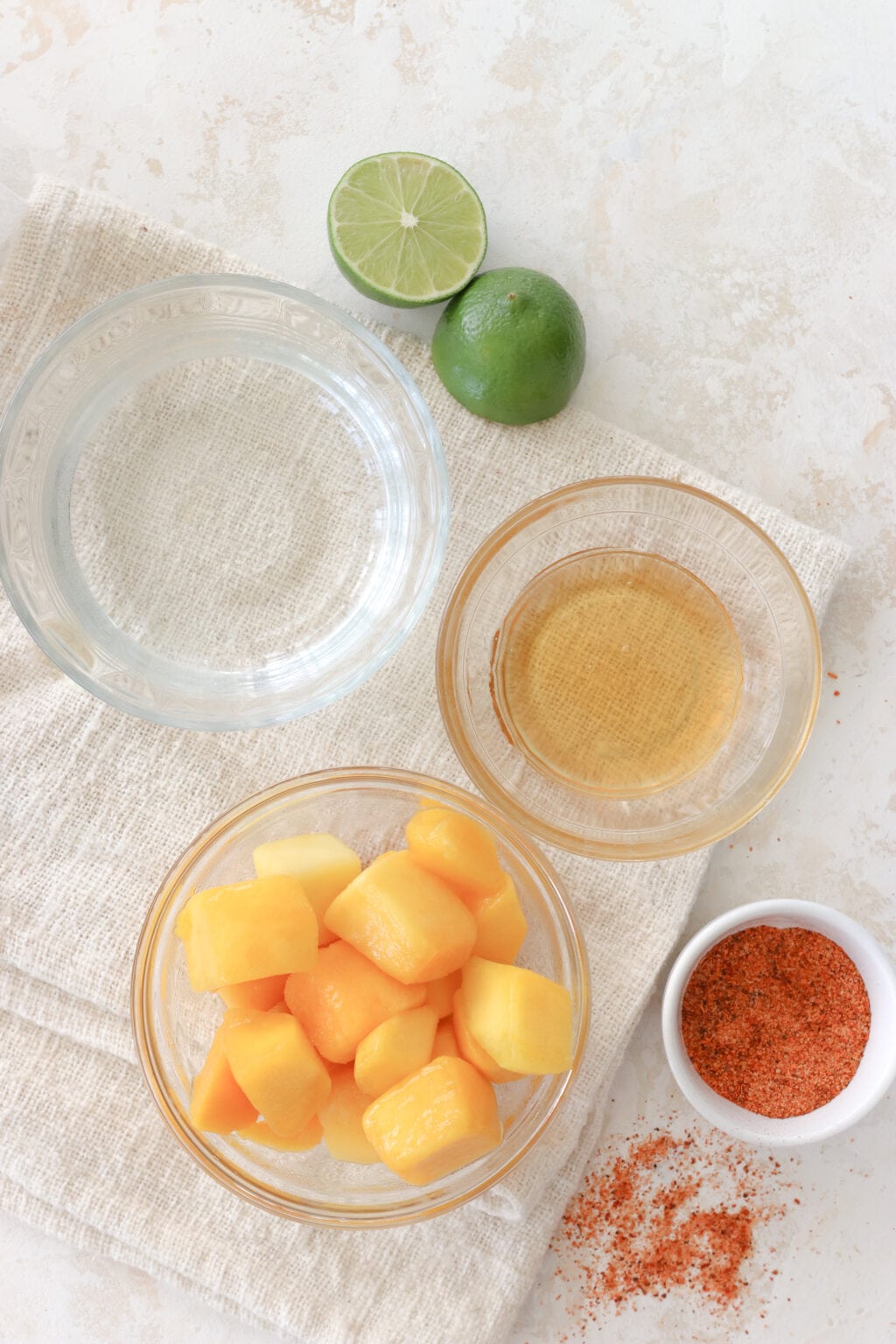 The ingredients to make mango tajin popsicles are on a white counter. There are limes cut in half, a bowl of water, a bowl of honey, a bowl of frozen mango and a small bowl of tajin