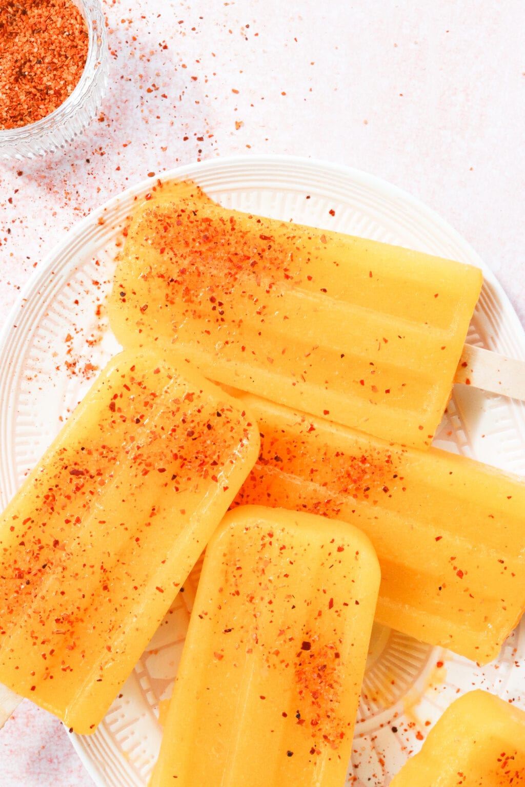 4 orange yellow coloured popsicles are on a pink and white oval plate. The popsicles are sprinkled with tajin seasoning.