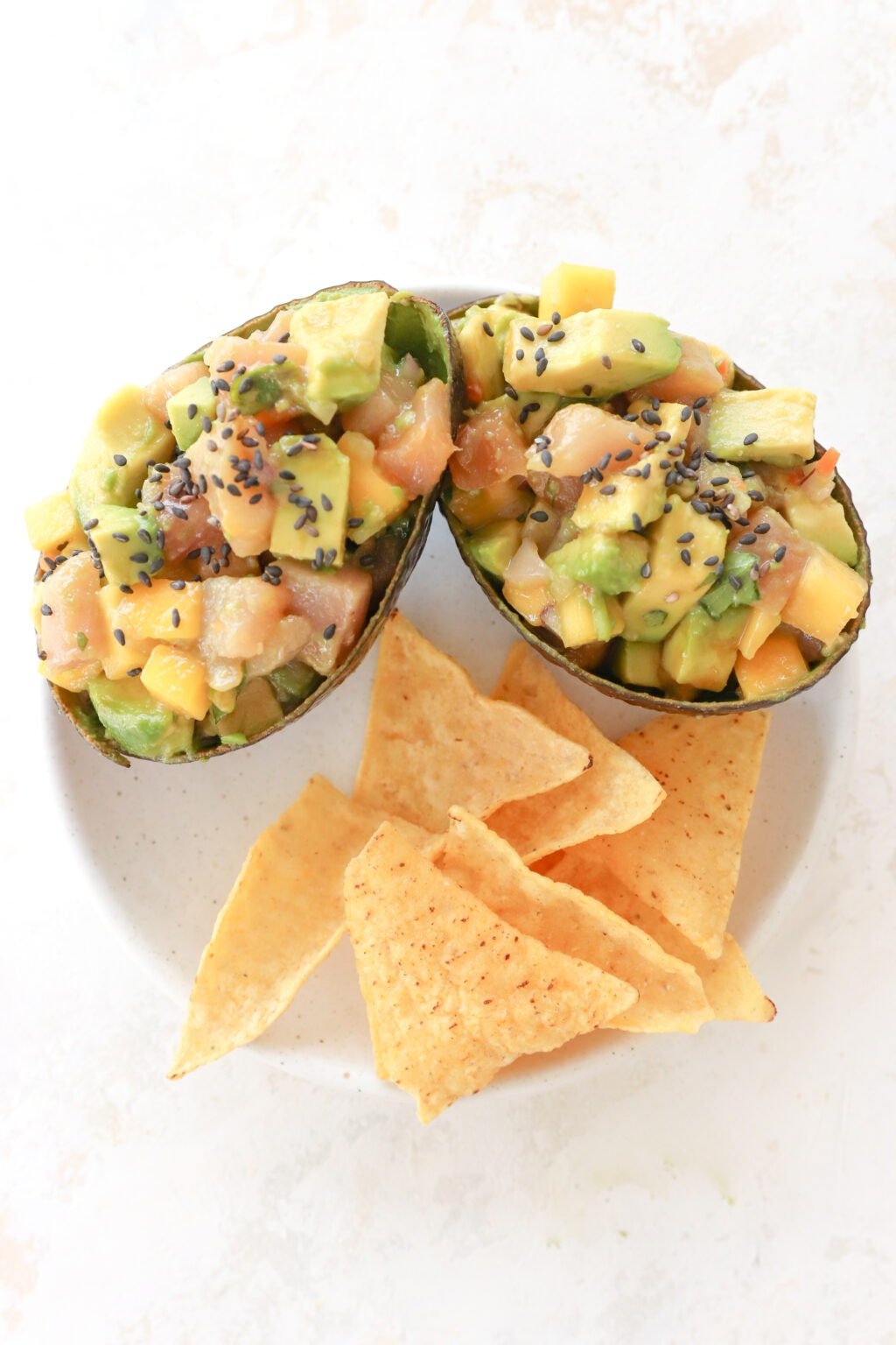 Two avocados are on a white plate. The avocados are stuffed with cubed avocado, cubed raw tuna, mango habanero salsa and topped with black sesame seeds. Below them on the plate are tortilla chips.