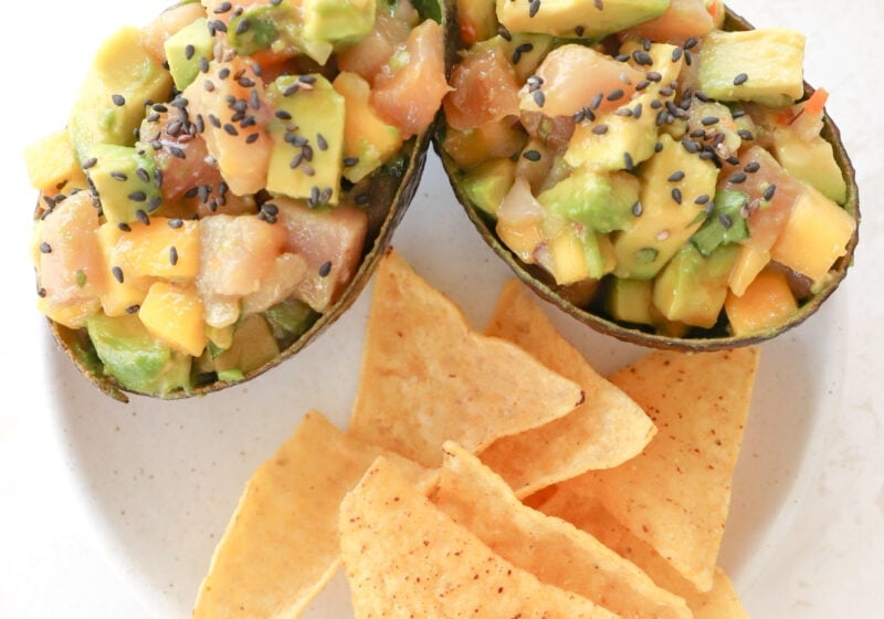 Two avocados are on a white plate. The avocados are stuffed with cubed avocado, cubed raw tuna, mango habanero salsa and topped with black sesame seeds. Below them on the plate are tortilla chips.