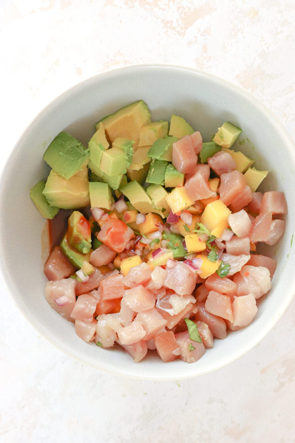A white bowl has the ingredients for tuna avocado boats in it. At the top is cubed avocado, then mango habanero salsa, then cubed sushi-grade tuna (which is pink), and then soy sauce.