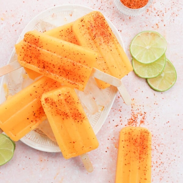4 popsicles orange yellow in color are on a white and pink plate they are covered with tajin seasoning and surrounded by lime rounds. There is a popsicle off the plate in the corner of the photo.