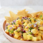 A pink plate with a rim is filled with mango habanero salsa and tortilla chips. The mango is cubed and mixed in with red onion and cilantro.