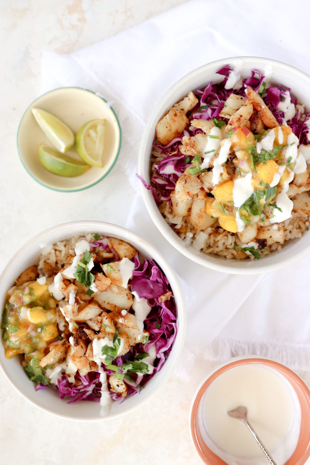 Two white bowls are the focus of the image. There is also a small bowl of lime slices in the lefthand top corned and a small bowl of white lime crema in the bottom right hand/ The bowls are filled with brown rice, mango and habanero salsa, purple cabbage, cajun cod, lime crema and cilantro.