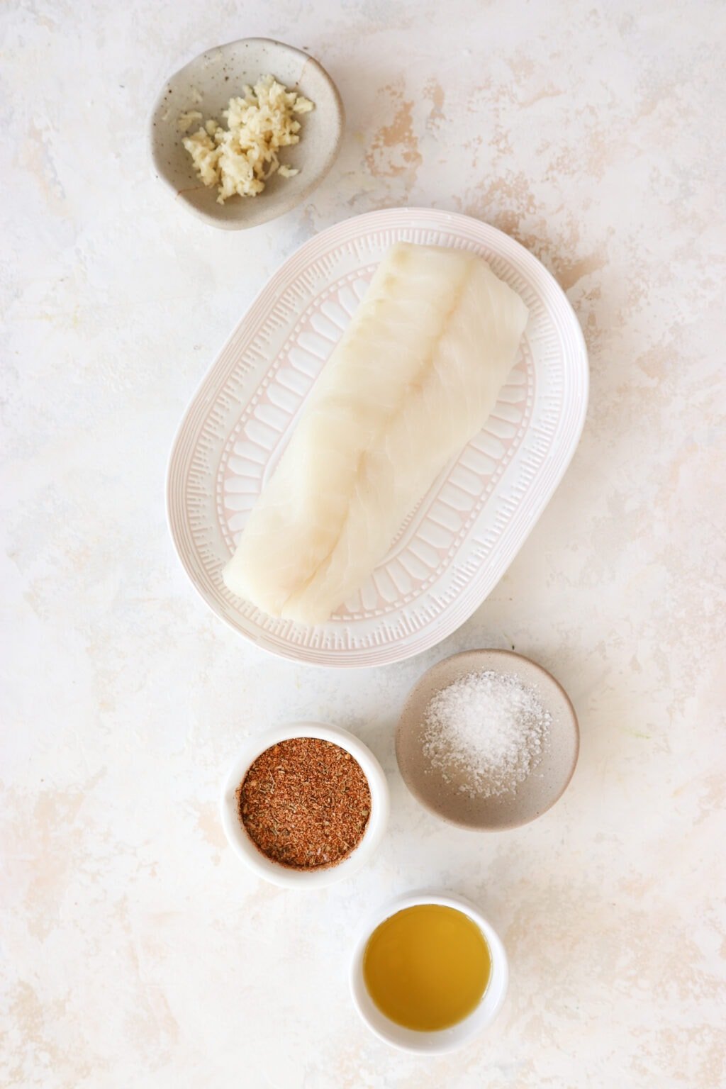 An overhead shot of ingredients on plates. At the top is a small white bowl with minced garlic, in the middle is a pink and white plate with white fish on it, and at the bottom are a grey bowl with salt in it and a white bowl with orangish brown cajun seasoning. 