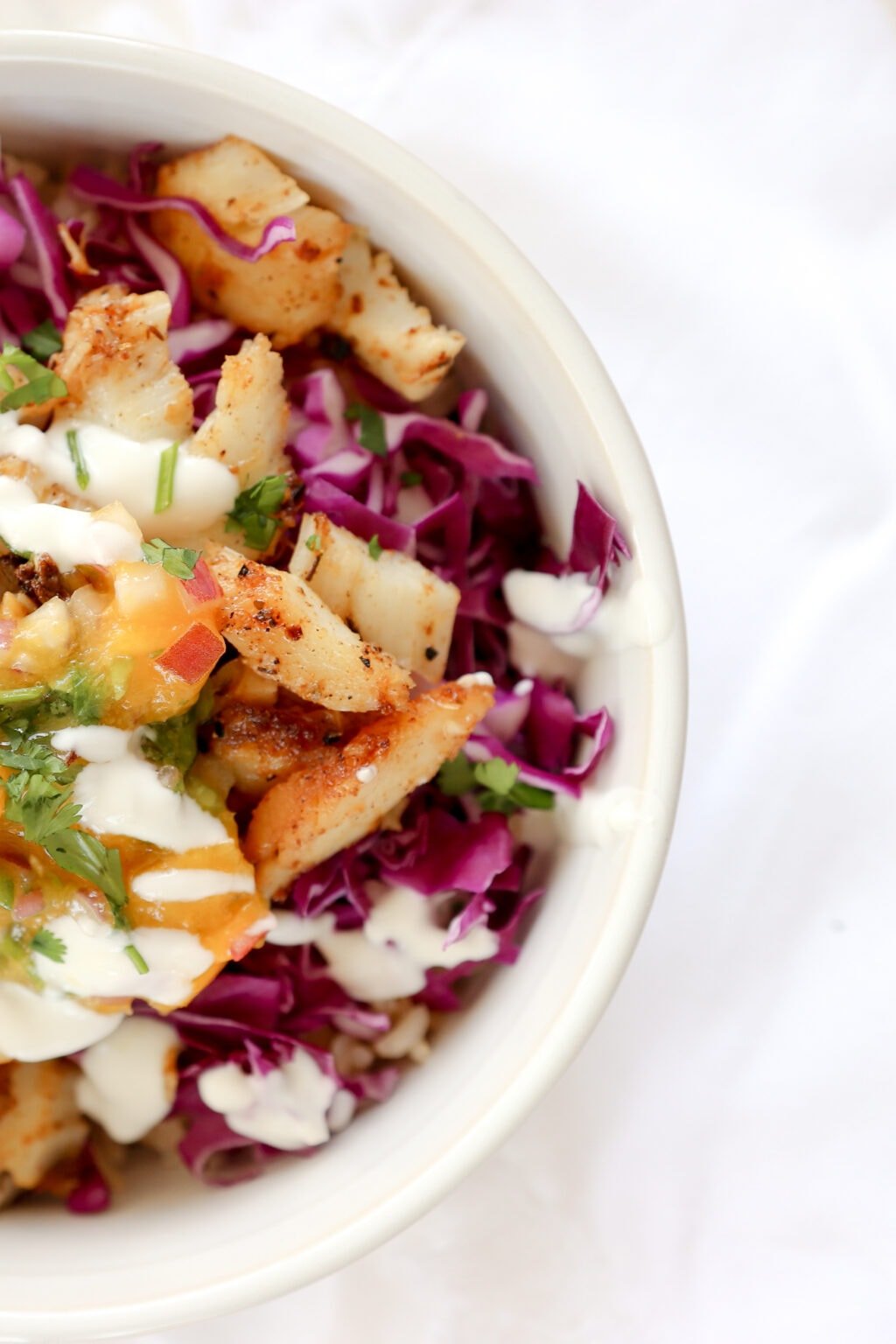 A zoomed in image of a white bowl with a fish taco bowl in it. There is shredded purple cabbage, cajun cod, green cilantro leaves, and white lime crema sauce. 