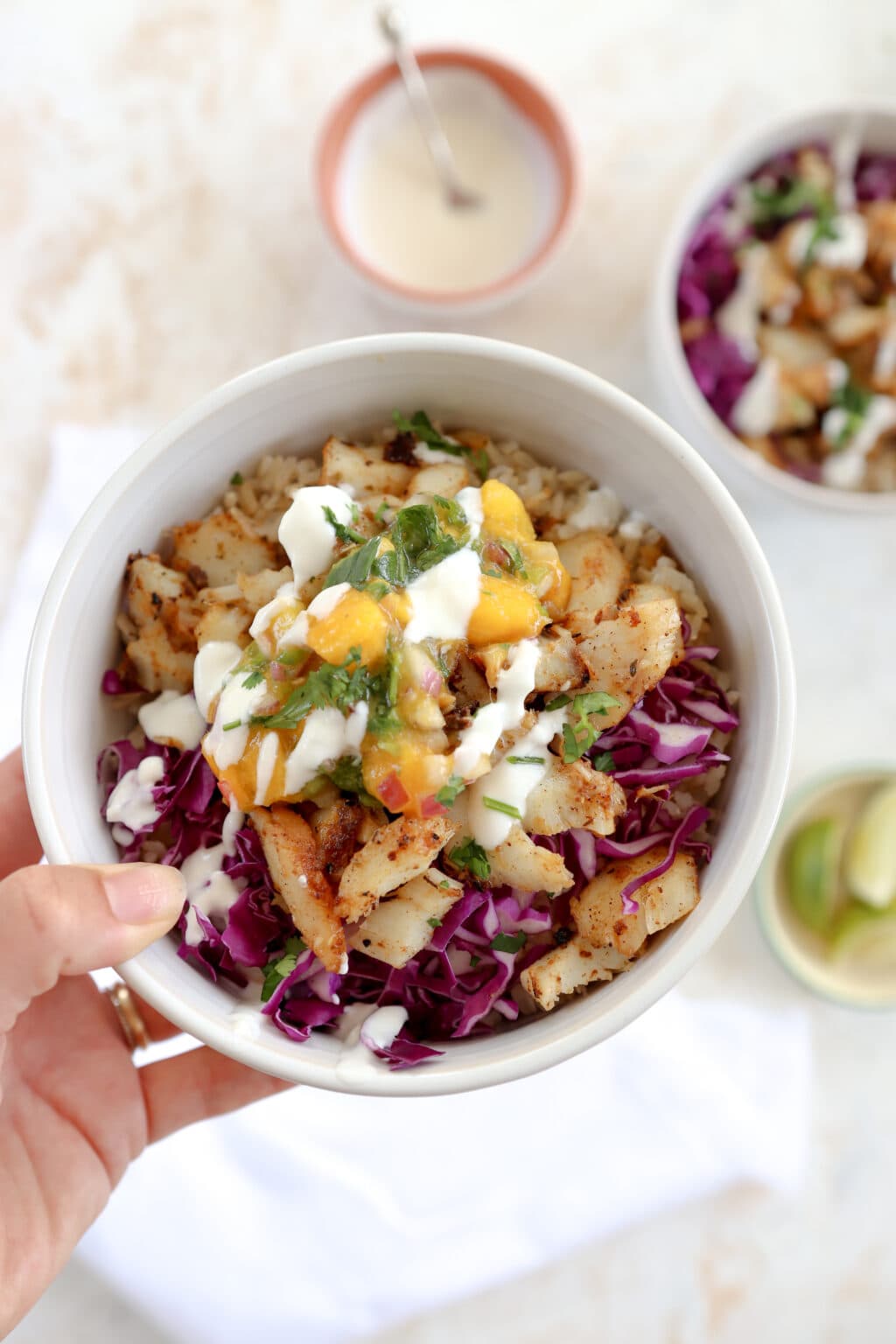 A hand is holding a white bowl. In the background are slices of lime, another taco bowl, and a bowl of lime crema. The main bowl in focus is filled with cabbage, cajun cod, lime crema, brown rice, mango, and cilantro. 