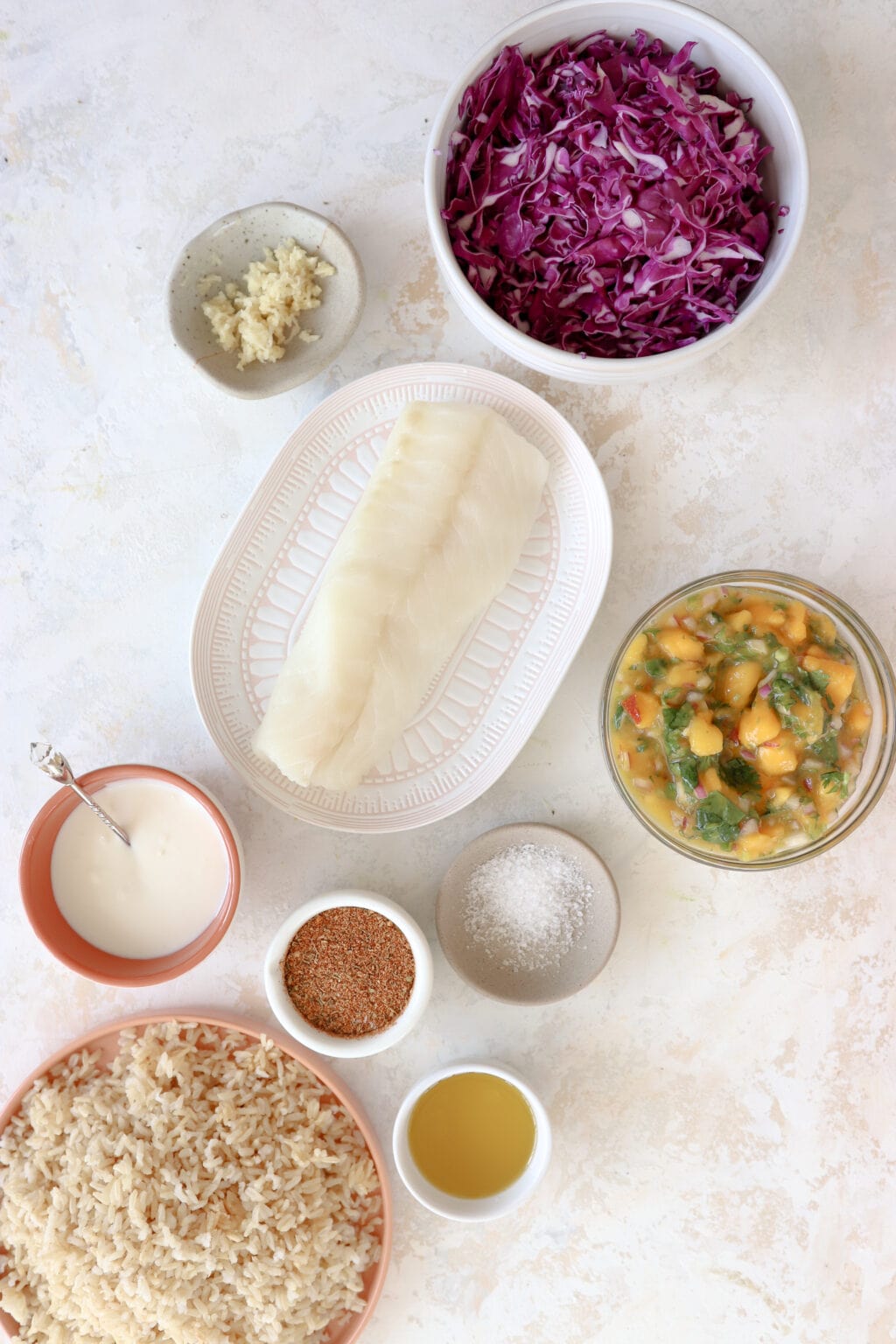 Overhead shot of ingredients for a cajun fish taco bowl recipe. In a 10 ounce white bowl is shredded purple cabbage, to the left is a 3 ounce bowl with minced garlic, to the right of that (in the center of the image) is a pink plate with raw white fish on it, to the right of that is a clear bowl with yellow and green salsa. Below the fish is a red ceramic bowl with lime crema and a spoon. Beside that is a 3 ounce white bowl with cajun seasoning and beside that is grey dish with salt. At the very bottom is a pink plate with brow rice on it and a 3 ounce white bowl with avocado oil.