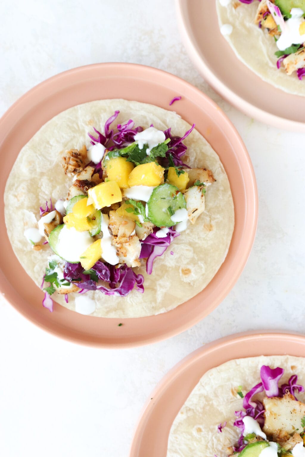 3 pink plates each have a tortilla with cajun fish tacos in them. Two of the plates are cut off in the right-hand upper and lower corner. The plate in the middle displays all the ingredients in the taco: purple cabbage, cilantro, mango, cajun cod, and lime crema. 