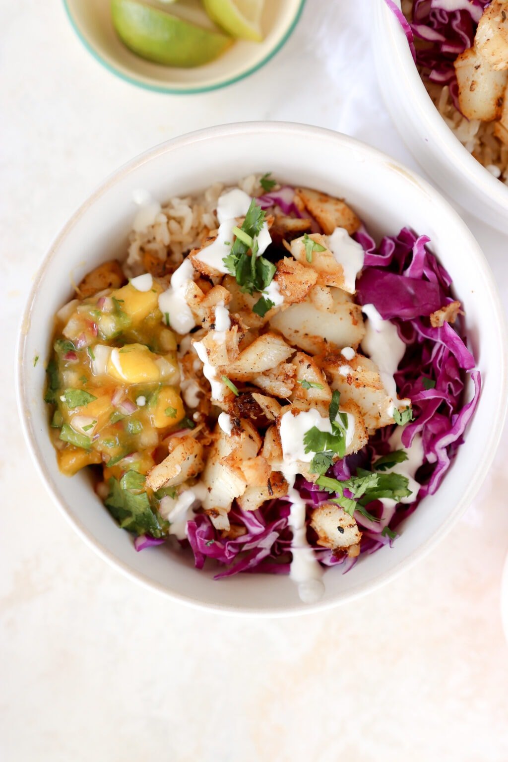 A white bowl is in the center of the photo. It is filled with brown rice, purple cabbage, mango habanero salsa, and cajun cod. The dish is topped with a drizzle of white sauce and cilantro.